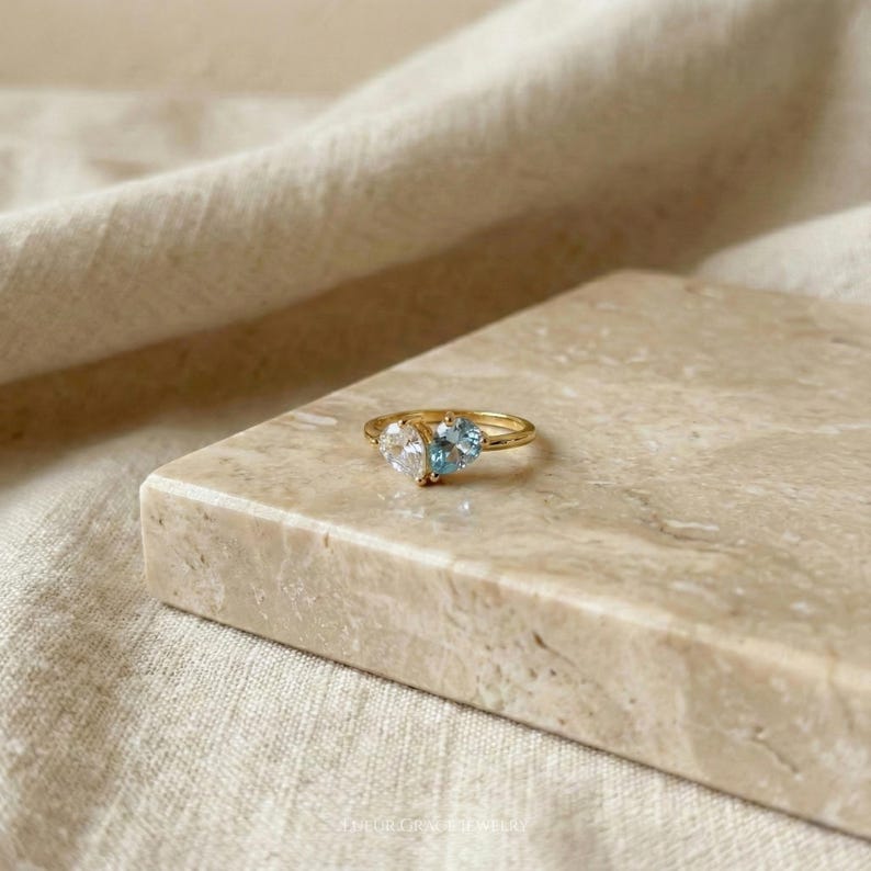 May include: A gold ring featuring two heart-shaped gemstones, one clear and one light blue. The ring is set on a marble surface with a neutral-toned fabric in the background. The ring is a piece of jewelry.