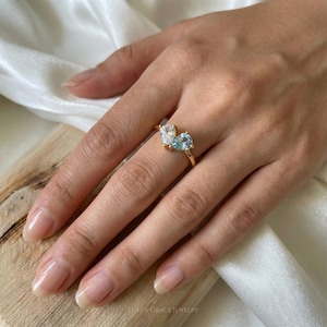 May include: A gold ring featuring two pear-shaped gemstones, one clear and one light blue. The ring is worn on a hand, with the gemstones positioned side by side. The ring is from LUFUR GRACE JEWELRY.
