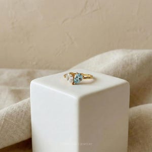 May include: A gold ring featuring two heart-shaped gemstones, one clear and one light blue. The ring is set on a white square block against a neutral background. The ring is a piece of jewelry.