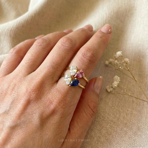 May include: Two gold rings with pear-shaped gemstones in white, pink, and blue. The rings are on a hand with a neutral background. The rings are delicate and elegant, perfect for everyday wear or special occasions.