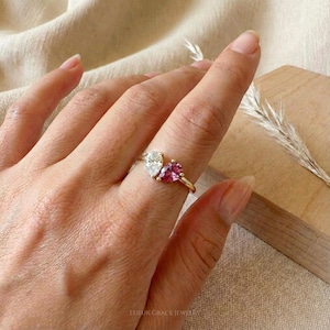 May include: A gold ring featuring two pear-shaped gemstones, one clear and one pink. The ring is on a finger, showcasing the jewelry's design and color contrast. The ring is a piece of fine jewelry.
