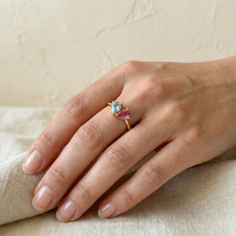 May include: A gold ring featuring two pear-shaped gemstones, one light blue and one pink. The ring is set on a delicate gold band. The gemstones are displayed on a hand.