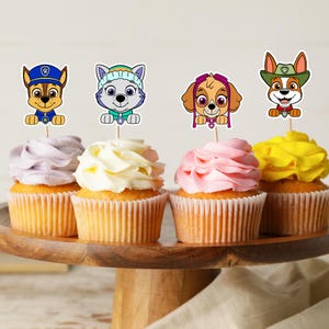 May include: Four cupcakes with colorful frosting and character toppers. The cupcakes sit on a wooden cake stand. The toppers feature cartoon dog characters in various outfits, including a police hat and a ranger hat. The frosting colors are lavender, white, pink, and yellow.
