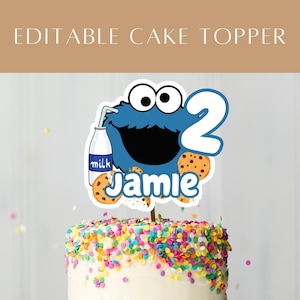 Könnte beinhalten: Ein Tortenaufsatz mit einer blauen Cartoonfigur und der Zahl 2. Die Figur hält eine Milchflasche und ist von Keksen umgeben. Der Name "Jamie" steht darunter. Der Kuchen ist mit bunten Streuseln verziert.