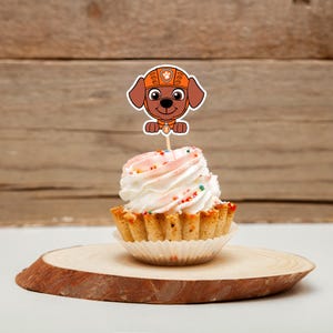 May include: A cupcake with white frosting, pink accents, and colorful sprinkles, topped with a cartoon dog cake topper. The cupcake sits on a wooden slice, with a wooden background.