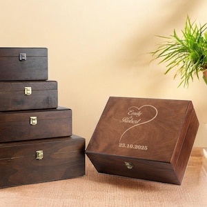 May include: A collection of wooden boxes in varying sizes, stained a dark brown. The boxes have metal clasps and hinges. One box has the names "Emily Robert" and the date "23.10.2025" engraved on the lid in a heart shape.