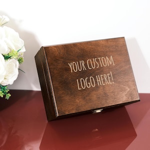 May include: A rectangular wooden box with a dark brown finish. The box has a hinged lid and a small metal clasp. The text "YOUR CUSTOM LOGO HERE!" is engraved on the top. White flowers are in the background.