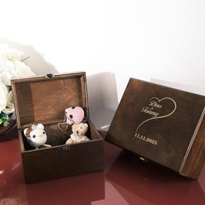 May include: Two dark wooden keepsake boxes, one open and one closed. The open box contains a crocheted cat, a teddy bear, and a pink keychain. The closed box has the names "Dina & Tommy" and the date "11.11.2025" engraved on the lid.