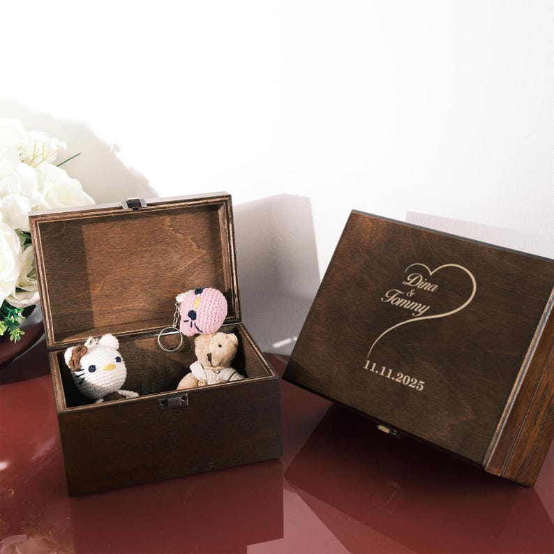 May include: Two dark wooden keepsake boxes, one open and one closed. The open box contains a crocheted cat, a teddy bear, and a pink keychain. The closed box has the names "Dina & Tommy" and the date "11.11.2025" engraved on the lid.