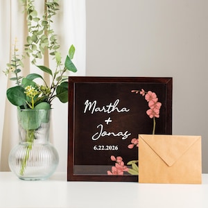 May include: A dark brown framed sign with the names "Martha + Jonas" and the date "6.22.2026" in white script, with pink floral accents. A kraft paper envelope rests beside the sign. A glass vase with greenery is in the background.