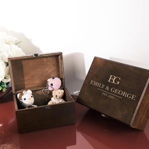 May include: Two wooden keepsake boxes, one open and one closed. The open box contains a crocheted cat, a teddy bear, and a pink keychain. The closed box is engraved with "EG" and "EMILY & GEORGE EST 2025".