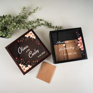 May include: Two shadow boxes with floral accents. One box is dark brown with the names "Olivia and Carlos" and the date "6.26.2026". The other box is black with the names "Maria Nicholas" and the date "July 25 2026". A brown envelope is in front of the boxes.