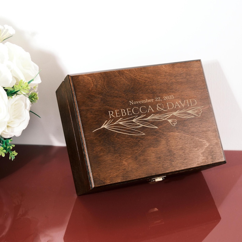 May include: A dark brown wooden box with a hinged lid. The lid is engraved with the names "REBECCA & DAVID" and the date "November 22, 2025", along with a floral design. The box is closed with a small metal clasp.