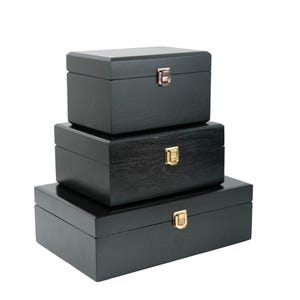 May include: Three black wooden boxes of different sizes, stacked. Each box is rectangular with a hinged lid and a metal clasp. The clasps are silver and gold in colour. The boxes are likely for storage.
