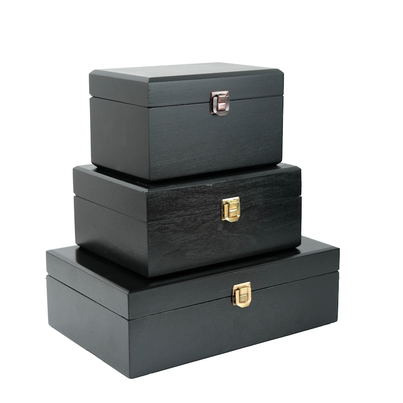 May include: Three black wooden boxes of different sizes, stacked. Each box is rectangular with a hinged lid and a metal clasp. The clasps are silver and gold in colour. The boxes are likely for storage.