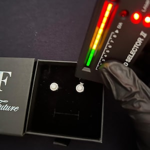 May include: A pair of round halo stud earrings with small white stones set in a black jewelry box. A Diamond Selector II testing device is positioned over the earrings, showing a green light indicator on its display panel.