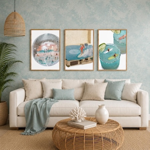 May include: Three framed wall art prints displayed above a beige sofa. The left print shows a disco ball filled with swimmers. The center print depicts a person surfing on a vinyl record player. The right print features two margaritas with surfers.
