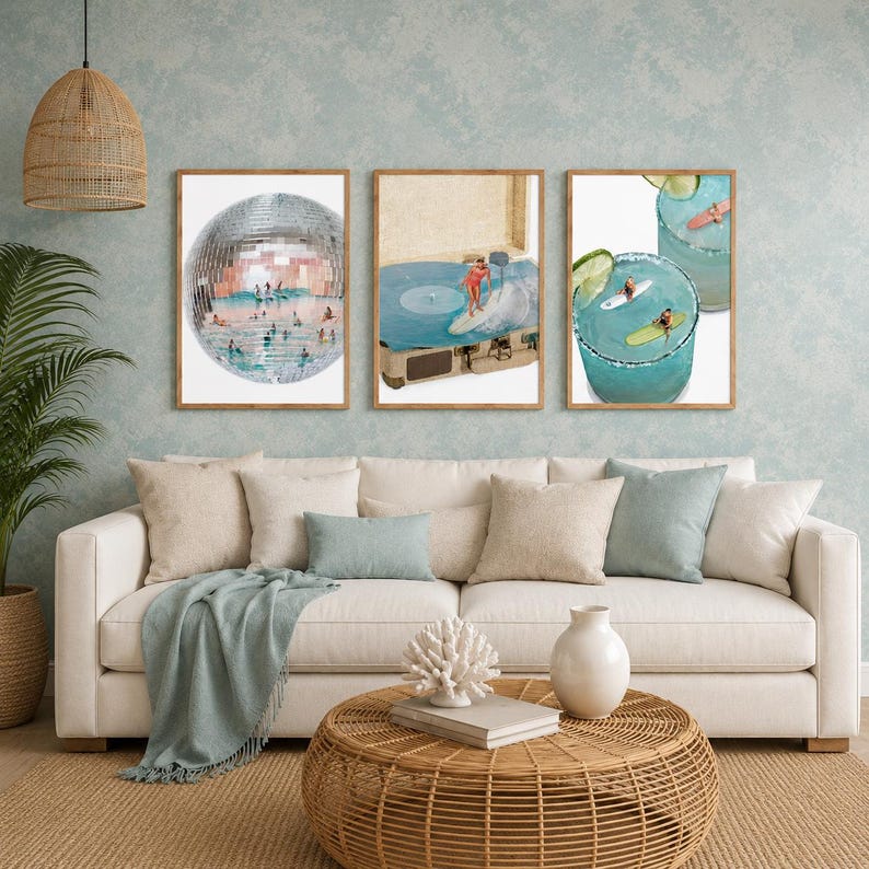 May include: Three framed wall art prints displayed above a beige sofa. The left print shows a disco ball filled with swimmers. The center print depicts a person surfing on a vinyl record player. The right print features two margaritas with surfers.