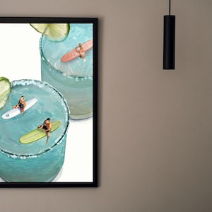 May include: A framed wall art print featuring two light blue cocktails in glasses with salt rims and lime slices. Inside the drinks, three miniature figures are surfing on small surfboards, with one person on a white board, one on a green board, and one on a pink board.