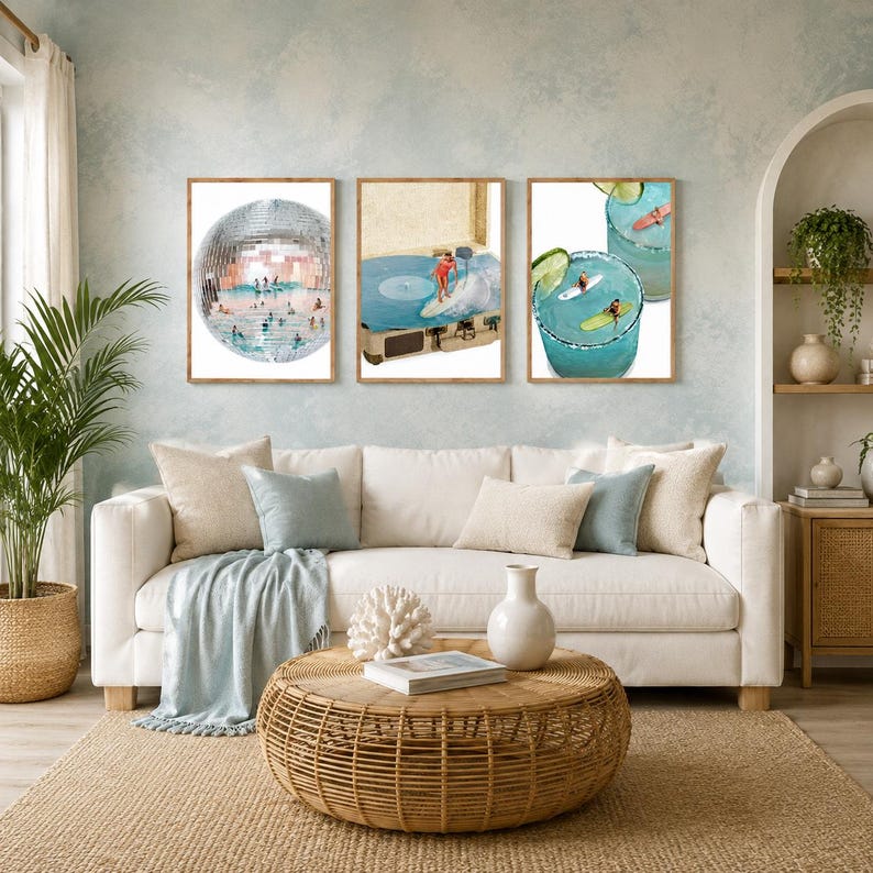 May include: Three framed wall art prints displayed above a sofa. The left print shows a disco ball filled with swimmers. The center print depicts a person surfing on a vinyl record. The right print features two glasses of blue cocktails with surfers.