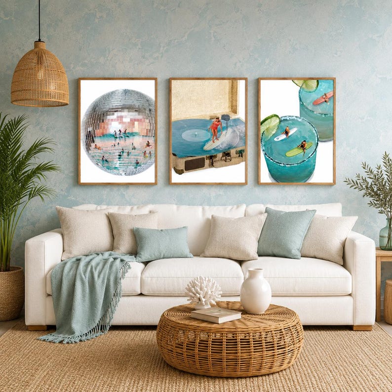 May include: Three framed wall art prints displayed above a white sofa. The left print shows a disco ball filled with swimmers. The center print features a record player with a surfer on the vinyl. The right print depicts two blue cocktails with surfers.