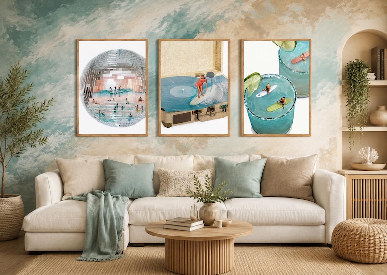 May include: Three framed wall art prints displayed above a white sofa. The left print shows a disco ball with people surfing inside. The center print features a record player with a surfer on the vinyl. The right print depicts two blue cocktails with surfers.