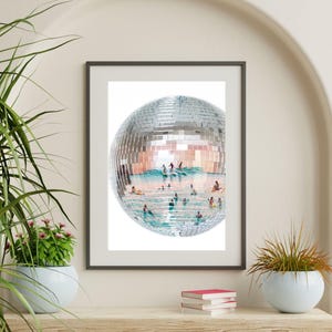 May include: A framed wall art print featuring a large, spherical disco ball composed of mirrored tiles. The surface of the disco ball reflects a scene of people swimming and surfing in turquoise ocean water with small waves under a bright sky.