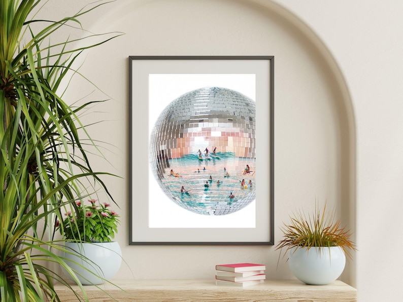 May include: A framed wall art print featuring a large, spherical disco ball composed of mirrored tiles. The surface of the disco ball reflects a scene of people swimming and surfing in turquoise ocean water with small waves under a bright sky.