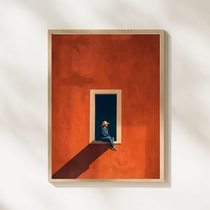 Könnte beinhalten: Gerahmter Kunstdruck mit einer Person, die in einem Fenster sitzt. Die Wand ist leuchtend orange, mit einem dunklen Schatten, der vom Fenster geworfen wird. Die Person trägt einen Hut und Jeanskleidung. Der Rahmen hat eine helle Holzfarbe.