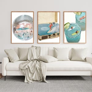 May include: Three framed art prints depicting a disco ball, a surfer on a record player, and two cocktails with tiny surfers. The prints are in wooden frames and hang above a white sofa with pillows and a blanket. A black lamp and a potted plant are also visible.