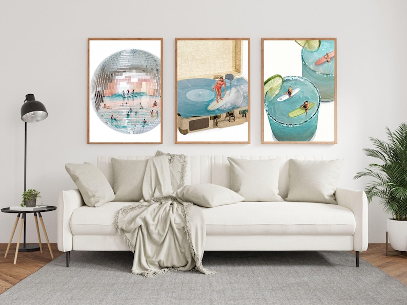 May include: Three framed wall art prints displayed above a white sofa. The left print shows a disco ball filled with swimmers. The center print features a person surfing on a vinyl record in a suitcase. The right print depicts surfers in cocktails.