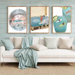 May include: Three framed wall art prints displayed above a white sofa. The left print shows a disco ball with surfers inside. The center print features a person surfing on a record player. The right print depicts surfers in blue cocktails.