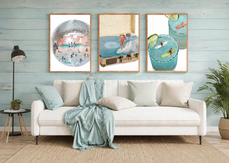 May include: Three framed wall art prints displayed above a white sofa. The left print shows a disco ball with surfers inside. The center print features a person surfing on a record player. The right print depicts surfers in blue cocktails.