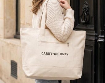 May include: A large, off-white canvas tote bag featuring two shoulder straps and a front exterior pocket with a zipper closure. The text CARRY-ON ONLY is printed in black, sans-serif capital letters on the front pocket of the bag.