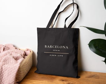 May include: Black tote bag with the word "BARCELONA" in white text, with "SPAIN" below it. Underneath is a line and the coordinates 41.39°N • 2.17°E. The bag has black handles.
