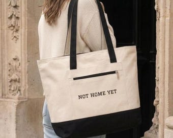 May include: A large cream-colored canvas tote bag with black shoulder straps and a black base. The front features a horizontal zippered pocket above the black text "NOT HOME YET" printed in a simple, sans-serif font centered on the bag's body.