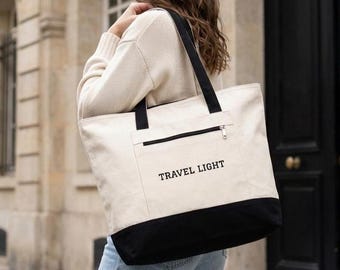 May include: A large canvas tote bag featuring a cream-colored upper section and a solid black base. The bag includes black shoulder straps, a front zippered pocket, and the text TRAVEL LIGHT printed in black on the front exterior panel.