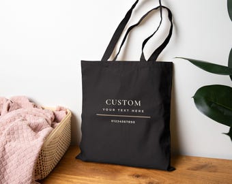 May include: A black tote bag with two shoulder straps featuring the white printed text CUSTOM YOUR TEXT HERE above a horizontal line and the number sequence 01234567890. The bag is made of fabric and has a rectangular shape with a flat base.