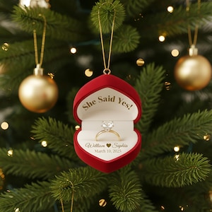 May include: A red heart-shaped Christmas ornament with the words "She Said Yes!" and a diamond ring inside. The ornament also includes the names "William ♥ Sophia" and the date "March 10, 2025".