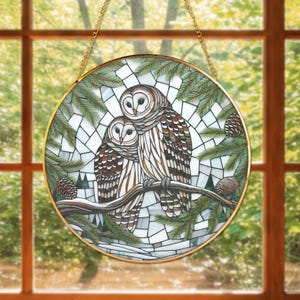 May include: A round stained glass window art piece featuring two owls perched on a branch. The owls have brown and white feathers, surrounded by pine needles and pine cones. The artwork is framed with a gold-colored metal and hangs from a gold chain.