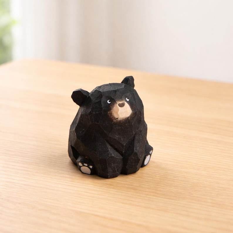 Pode incluir: Estatueta de urso de madeira preta com design geom&eacute;trico. O urso est&aacute; sentado com as patas vis&iacute;veis e tem um focinho de cor clara e pequenos detalhes brancos para os olhos. A estatueta est&aacute; sobre uma superf&iacute;cie de madeira clara.