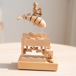 Puede incluir: Un autómata mecánico de madera clara que presenta un pez segmentado con una pequeña figura montada encima. El pez está suspendido sobre una base con forma de olas con un mecanismo de manivela, decorado con pequeños recortes de madera.