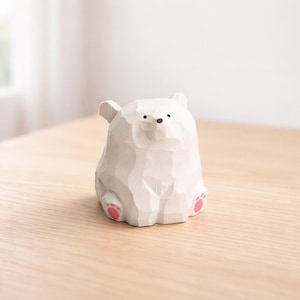 Pode incluir: Uma figura de urso polar de madeira branca esculpida com almofadas de pata rosa. O urso tem uma superf&iacute;cie texturizada e pequenos olhos e nariz pretos. A figura est&aacute; em uma superf&iacute;cie de madeira clara.