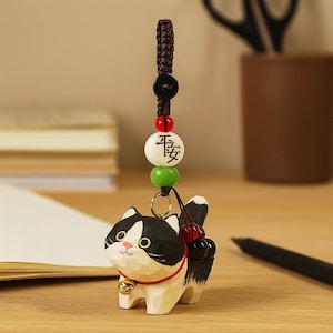 Peut inclure: Figurine de chat en bois sculpté, avec des marques noires et blanches, un collier rouge et une petite cloche dorée. Le chat est attaché à une chaîne de perles avec un bouton noir, des perles rouges et vertes et une perle blanche avec des caractères asiatiques.