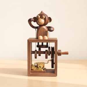 May include: A wooden automaton featuring a monkey figure with a dark brown body and light brown face, hands, and feet. The monkey sits atop a rectangular wooden frame containing a mechanical gear system, a hand crank, and a brass music box mechanism.