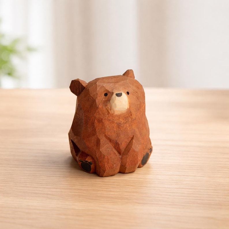 May include: A small, handcrafted wooden bear figurine. The bear is carved from brown wood with a light-colored snout and black eyes and paws. The bear is sitting down, and the wood grain is visible.