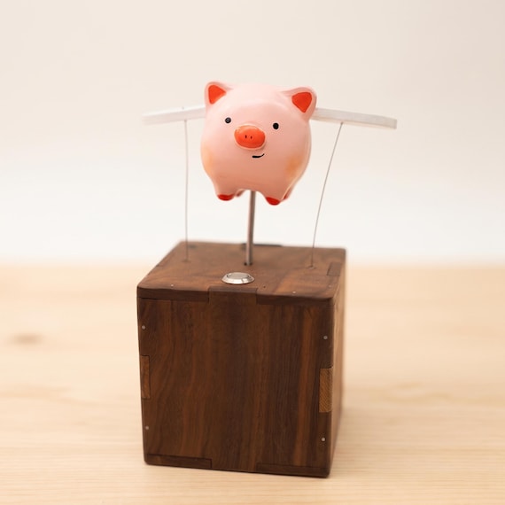 A battery-powered wooden automaton of a flying pig — the kind of whimsical desk companion that stops visitors mid-sentence to ask 'wait, what IS that?'