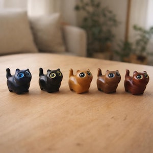 May include: A row of five small wooden cat figurines in varying shades of brown. The cats have large, expressive eyes, with colors including blue, yellow, and brown. Each cat is carved with a simple, rounded design, giving them a charming, whimsical appearance.