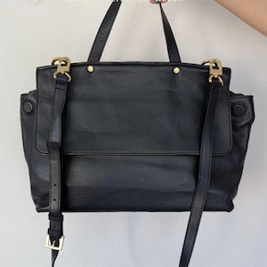 Puede incluir: Bolso de mano de cuero negro con herrajes dorados. El bolso tiene un asa superior, una correa para el hombro desmontable y un cierre de solapa. El bolso tiene un acabado texturizado y está sostenido por una mano. El bolso tiene el texto "Opalo ES" y "Casa de Joyeria "Tar Roban"" en el fondo.