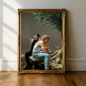 Puede incluir: Espejo enmarcado en oro con dos personas sentadas en un banco de piedra bajo un árbol. Una persona viste una camiseta sin mangas negra, la otra una camiseta sin mangas blanca y vaqueros azules. La escena tiene una cualidad natural y etérea.